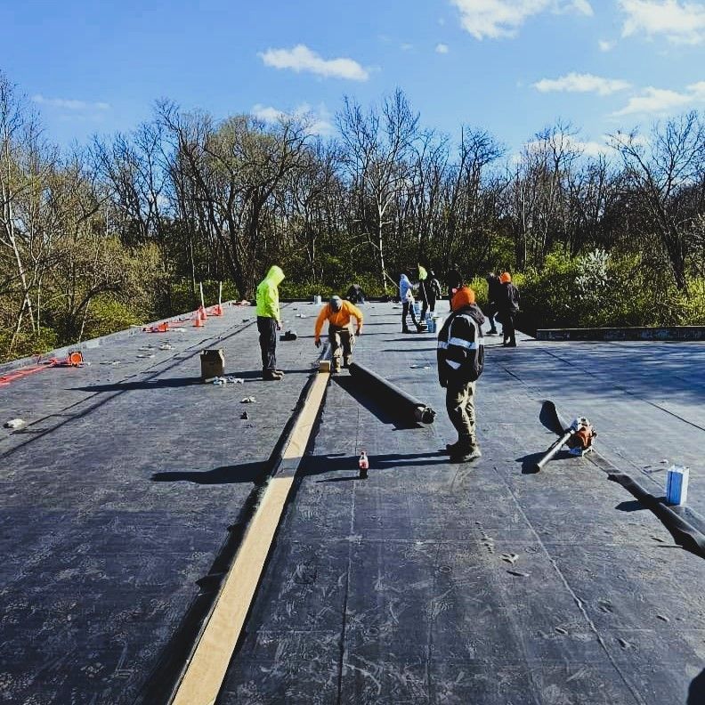 Workers installing a black roof membrane on a flat rooftop under a clear blue sky.