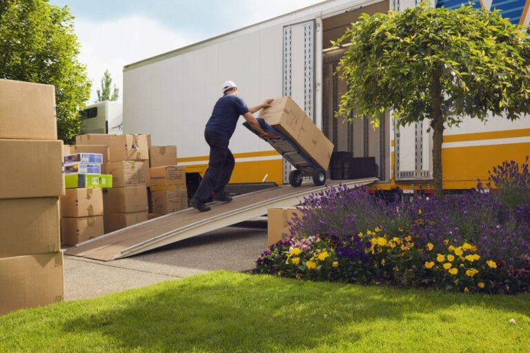 Person pushing a box on a hand truck up a ramp into a moving truck. Boxes stacked on lawn.