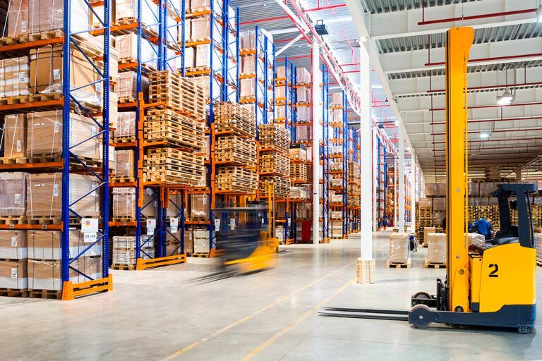 Warehouse interior with tall shelving packed with goods, two yellow forklifts.