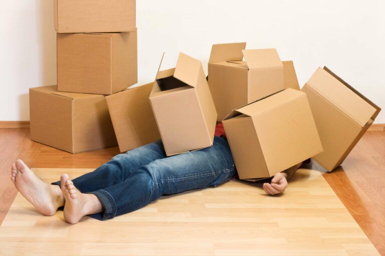 Person lying under a pile of cardboard moving boxes on a wooden floor.