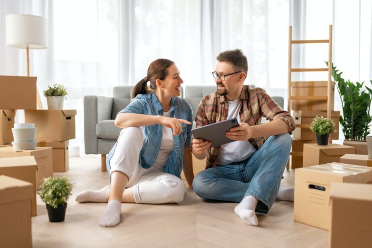Couple sitting on floor, looking at tablet, surrounded by moving boxes in new home.