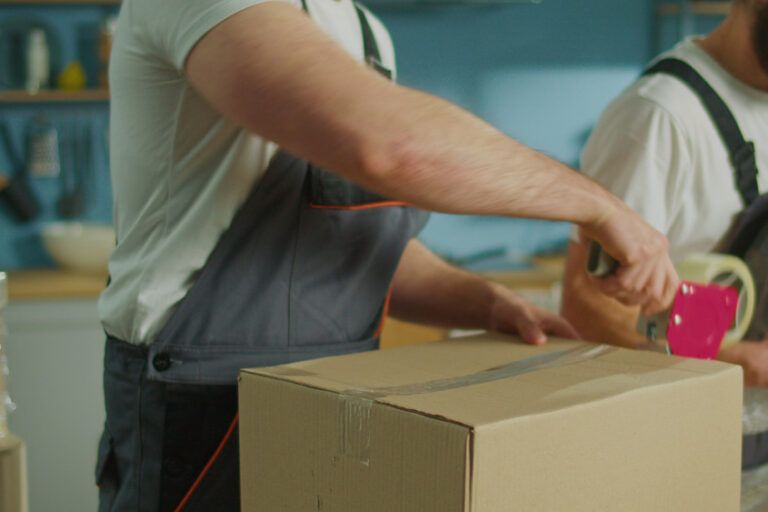Movers taping a cardboard box in a kitchen.