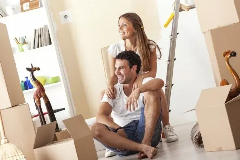 Couple smiling, sitting among moving boxes in a new home, looking content.