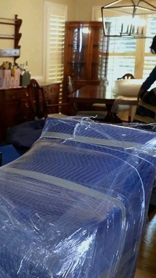 Furniture wrapped in blue padding and plastic, in a room with other furniture and a person.