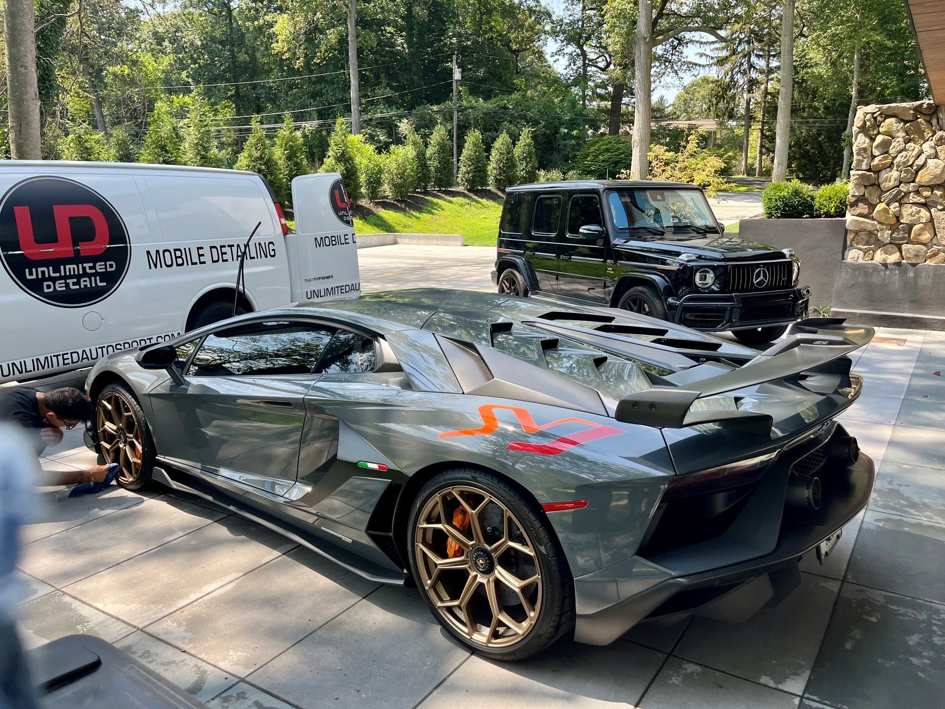 Gray sports car with bronze wheels parked beside a black SUV and white van on a sunny driveway