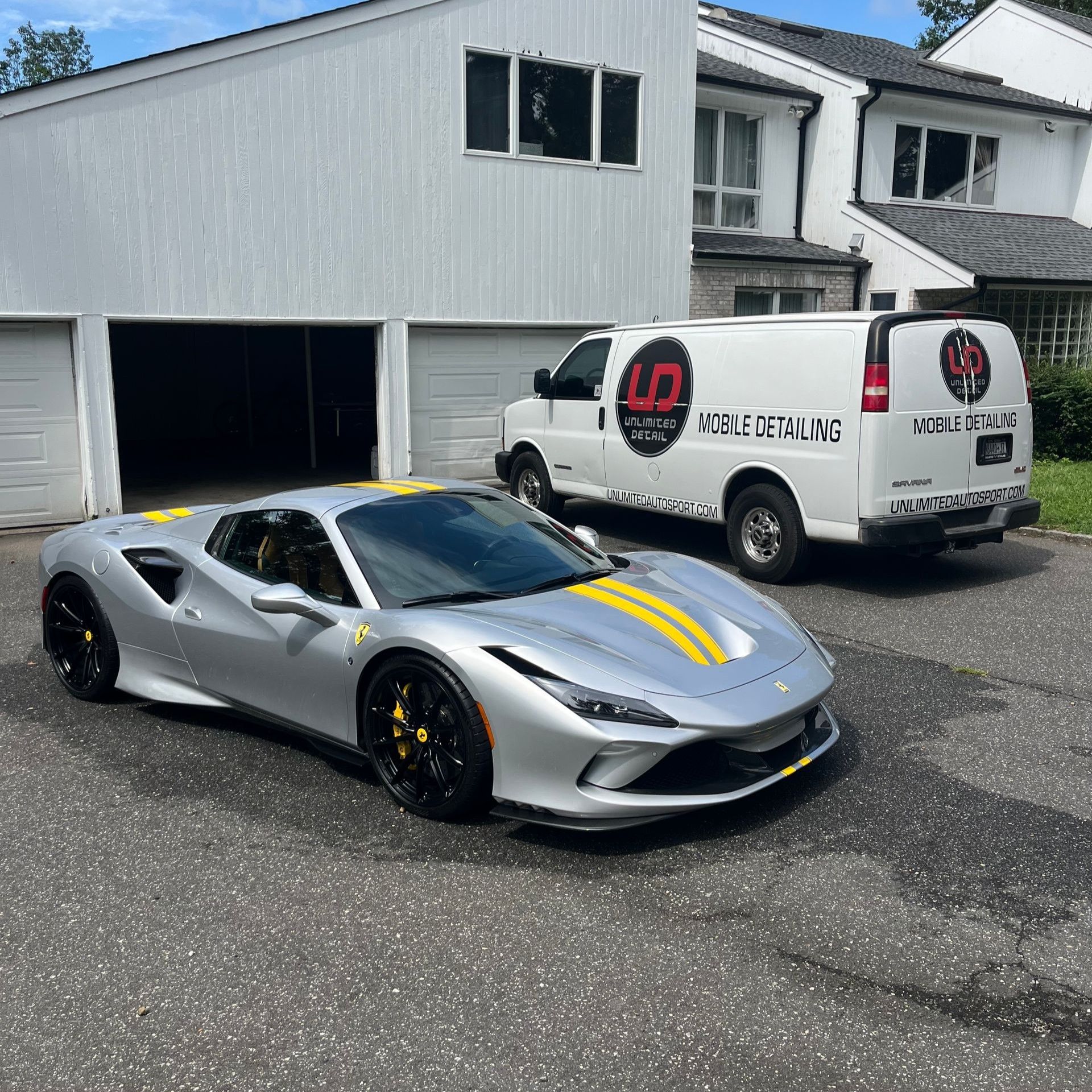 Silver sports car with yellow stripes parked beside a white service van in front of a garage.