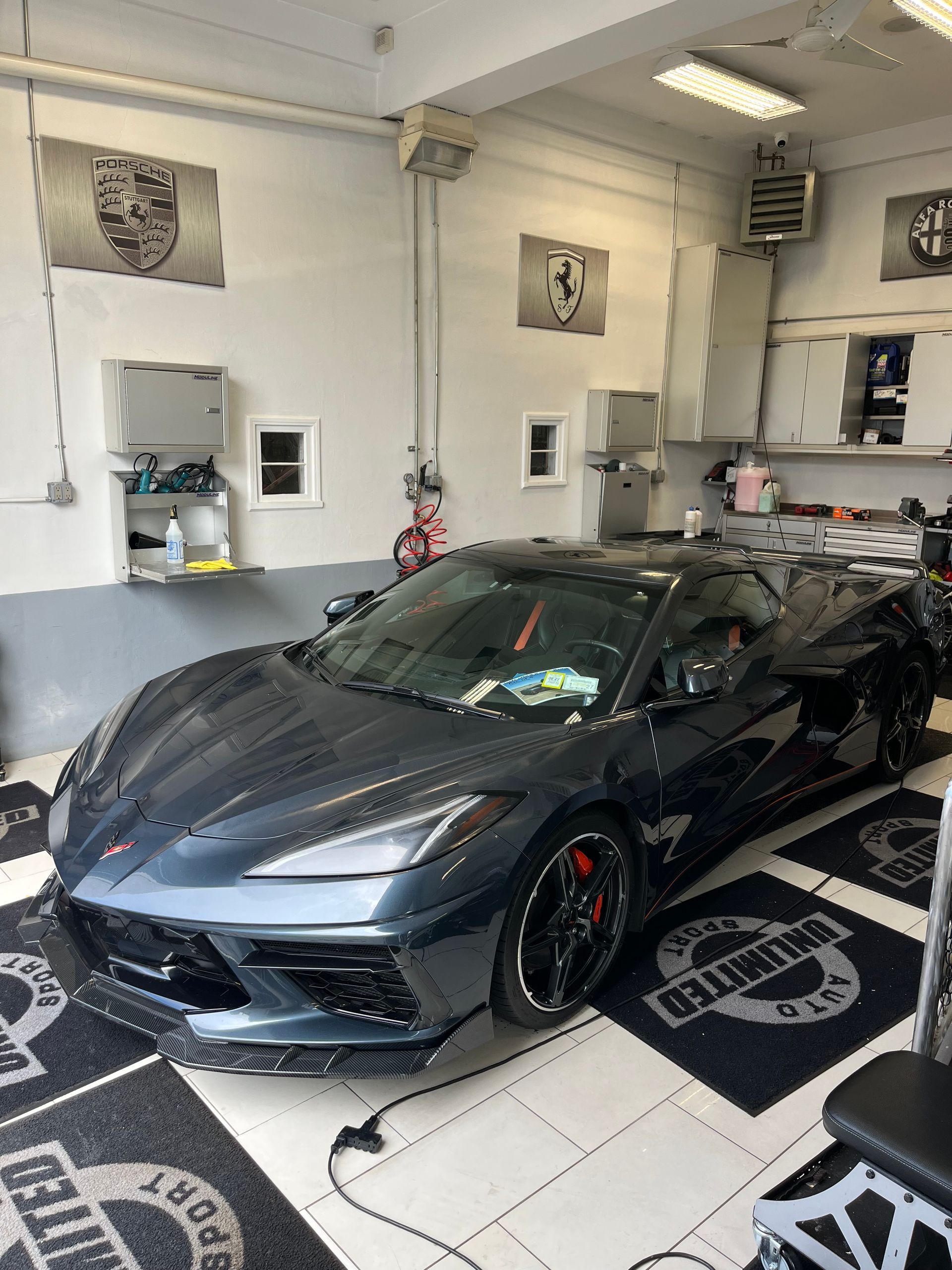 Dark gray sports car in a garage-like showroom, with white walls and black floor mats.