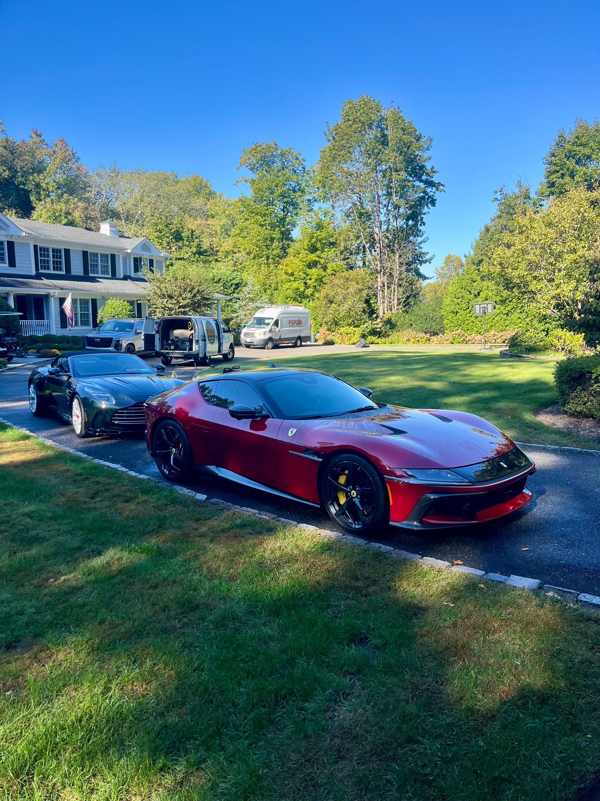 Red sports car parked on a driveway in front of a white house, with a black car behind it.