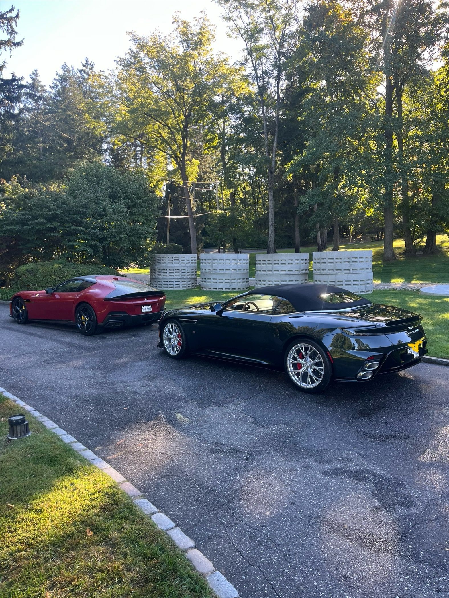 Two sports cars, a red and a black convertible, parked on a gravel driveway by trees and a white fence.