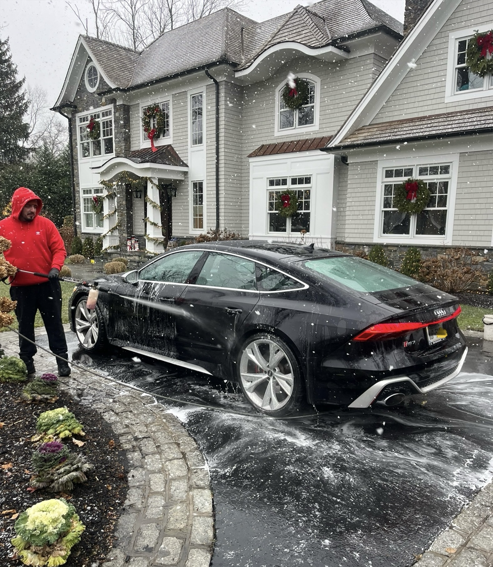 A man is washing a black car in front of a large house.