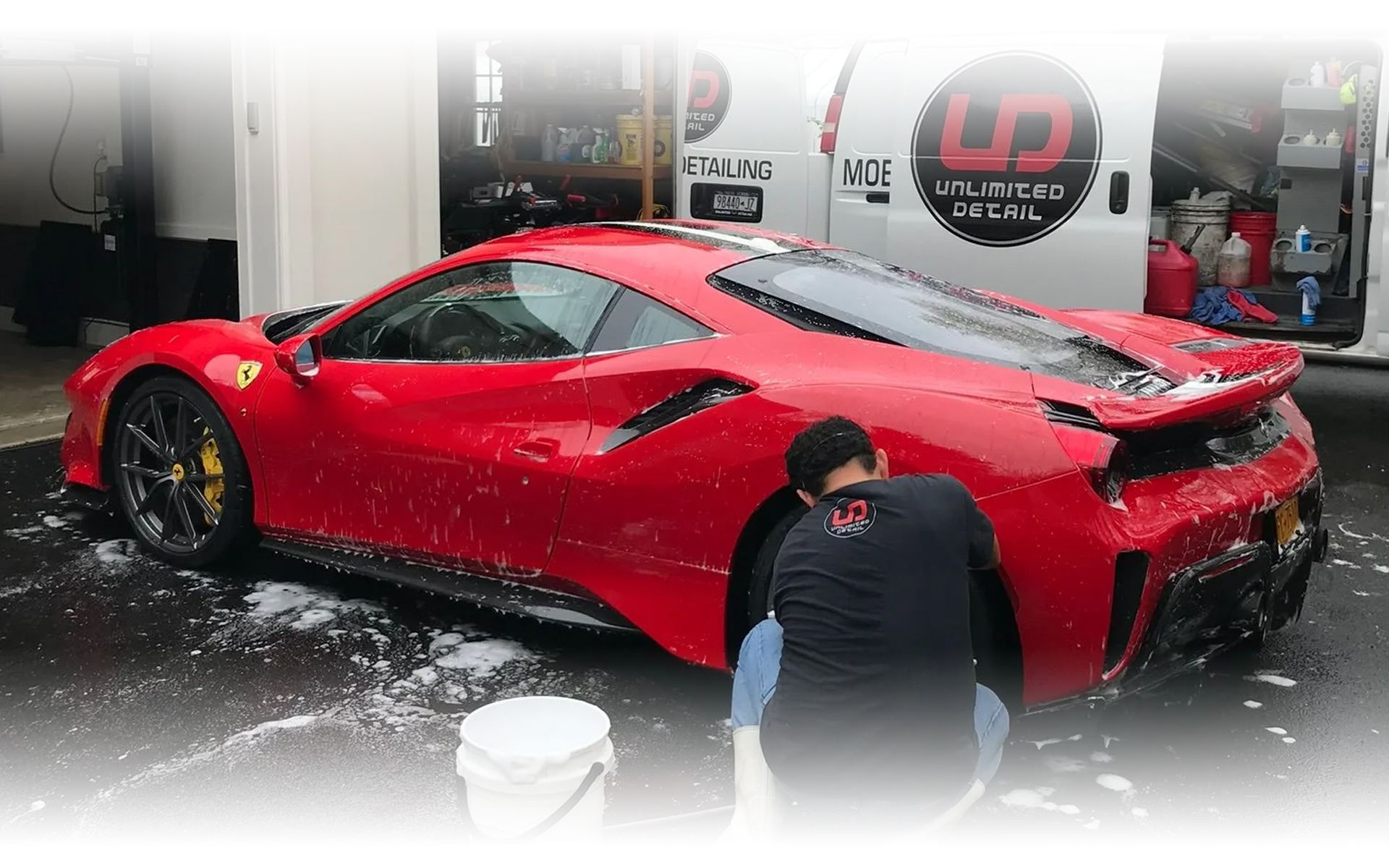 A man is washing a red sports car in front of a van.