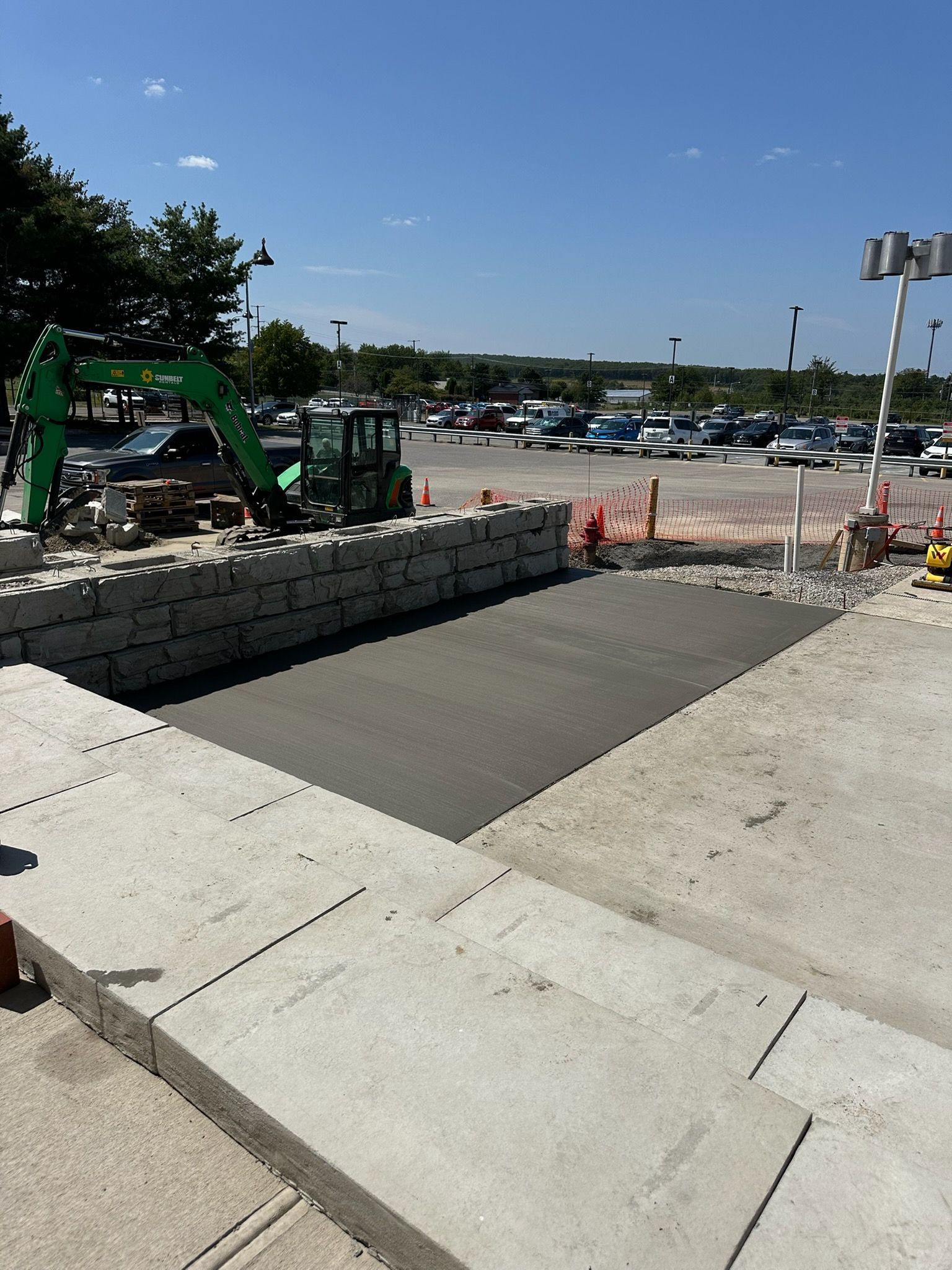 Construction site with a concrete ramp, stone retaining wall, and green excavator under a clear blue sky