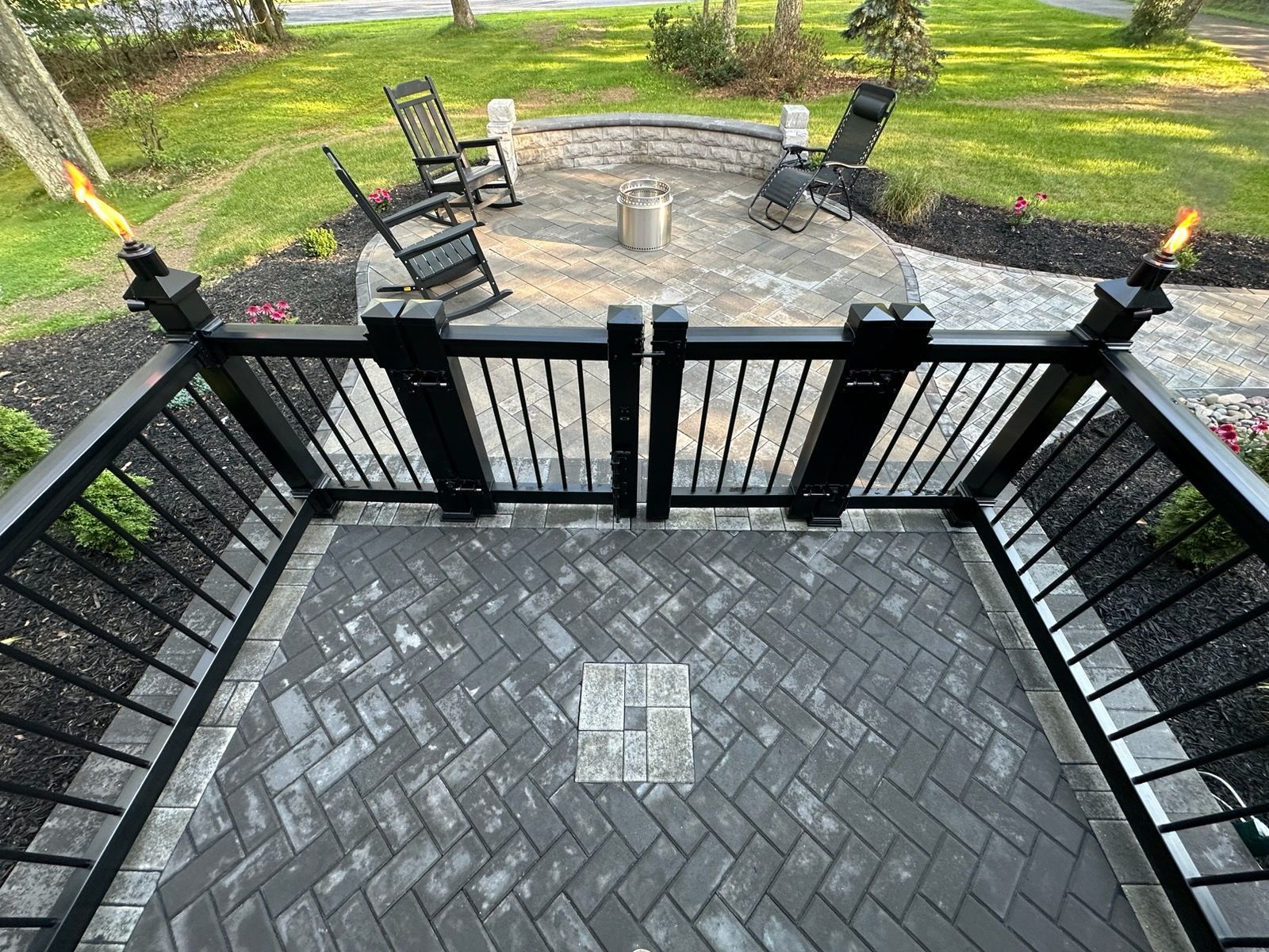 Black fence gate leading to a paved patio with chairs and a fire pit in a green backyard.