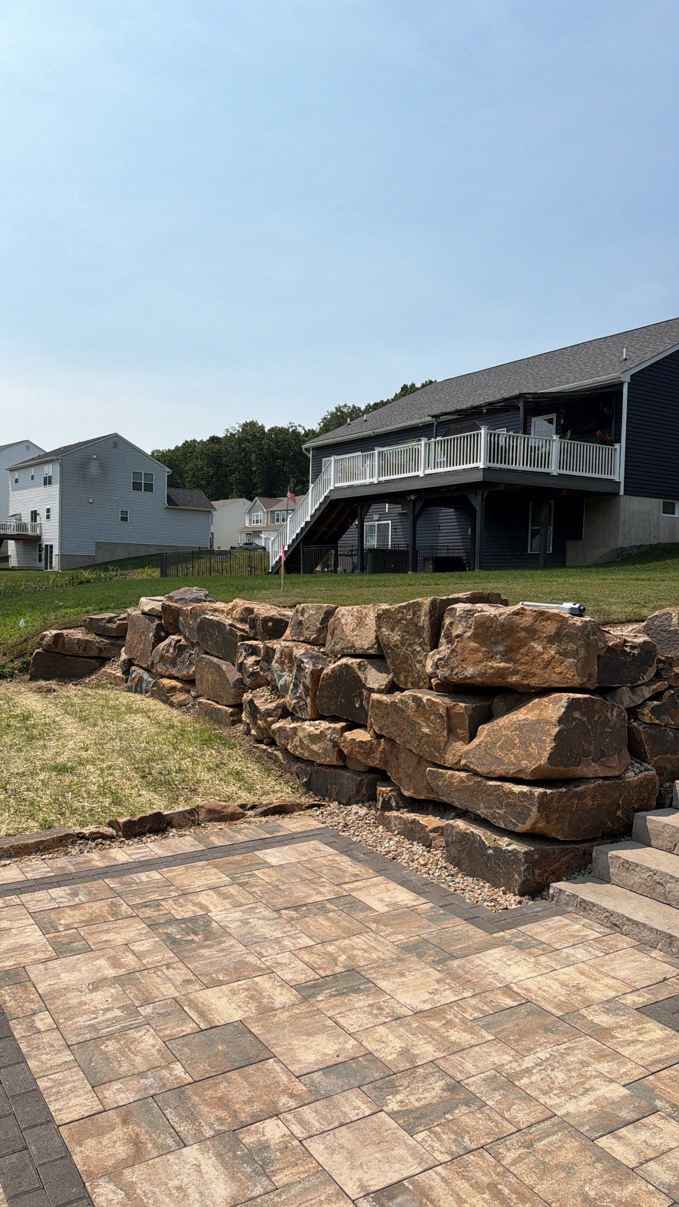 Stone retaining wall and tiled patio in front of a house with an elevated porch and sloped roof under a clear sky