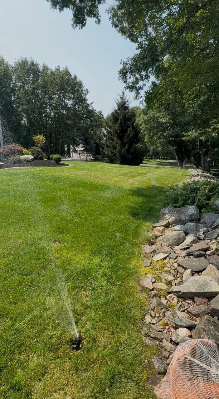 Lawn sprinkler watering a green yard beside a stone border and trees on a sunny day