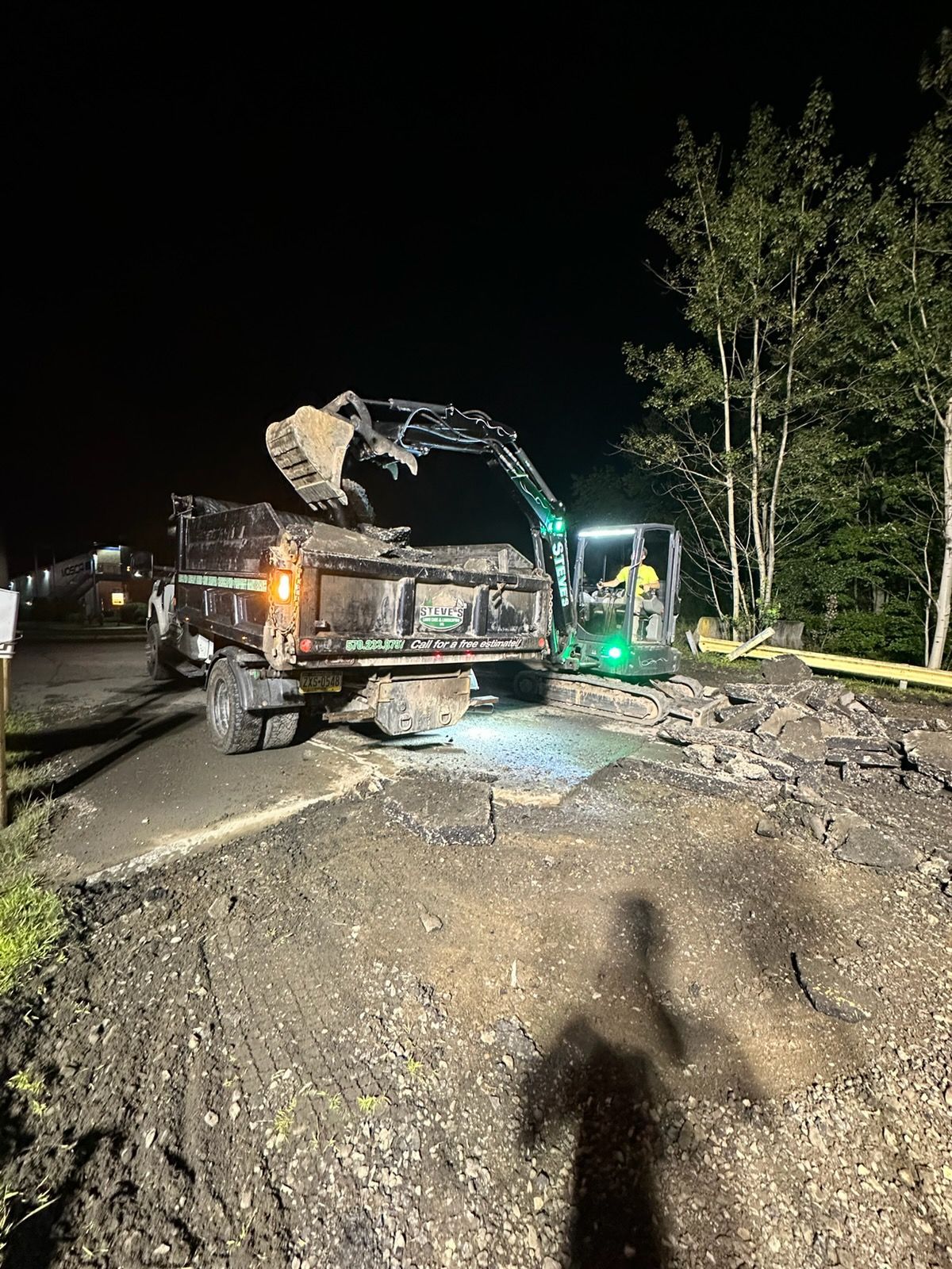 Nighttime roadwork with excavator loading debris into a dump truck beside a damaged road and trees