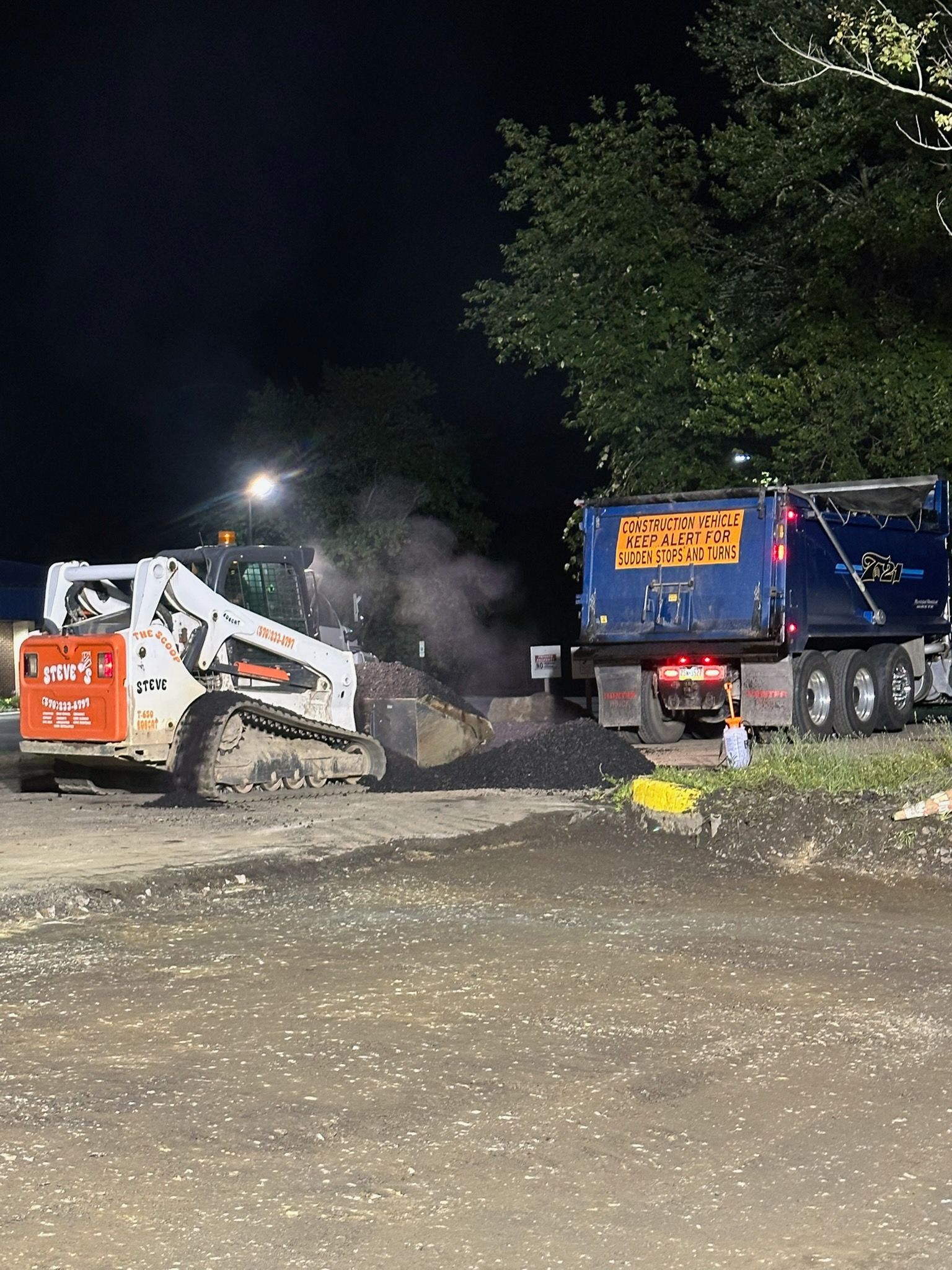 Construction vehicles at night, including a white skid steer and blue dump truck, on a gravel site.