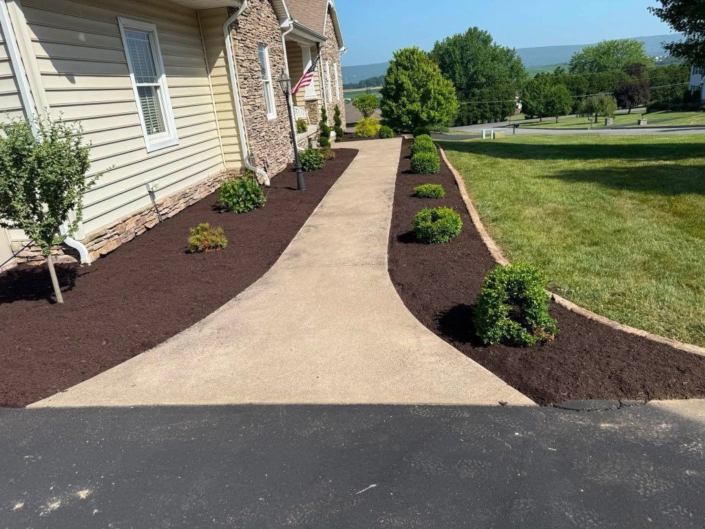 Walkway leading to a house entrance, bordered by dark mulch and small shrubs, with a lawn and hills beyond.