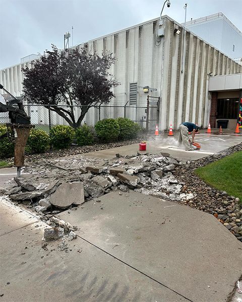 Concrete sidewalk being demolished outside a building. A worker operates a jackhammer.