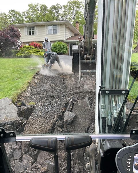 Workers using excavator to remove asphalt driveway in front of a house. One worker is breaking up asphalt with a tool.