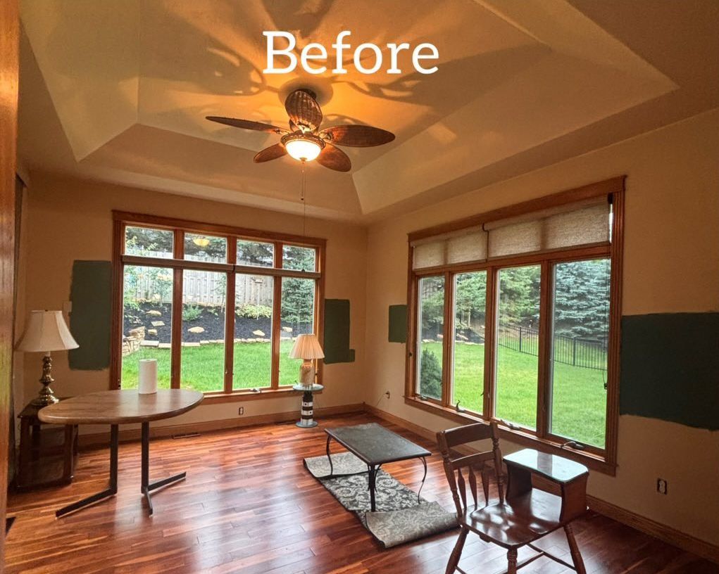 A room with tan walls, wood trim, large windows, a desk, and a ceiling fan. - before