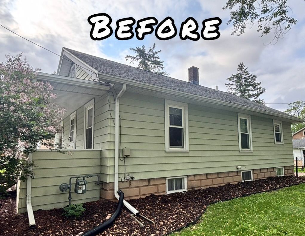 Green-sided house with white trim, before a remodel. Dark roof, brick foundation, and mulch bed. - before