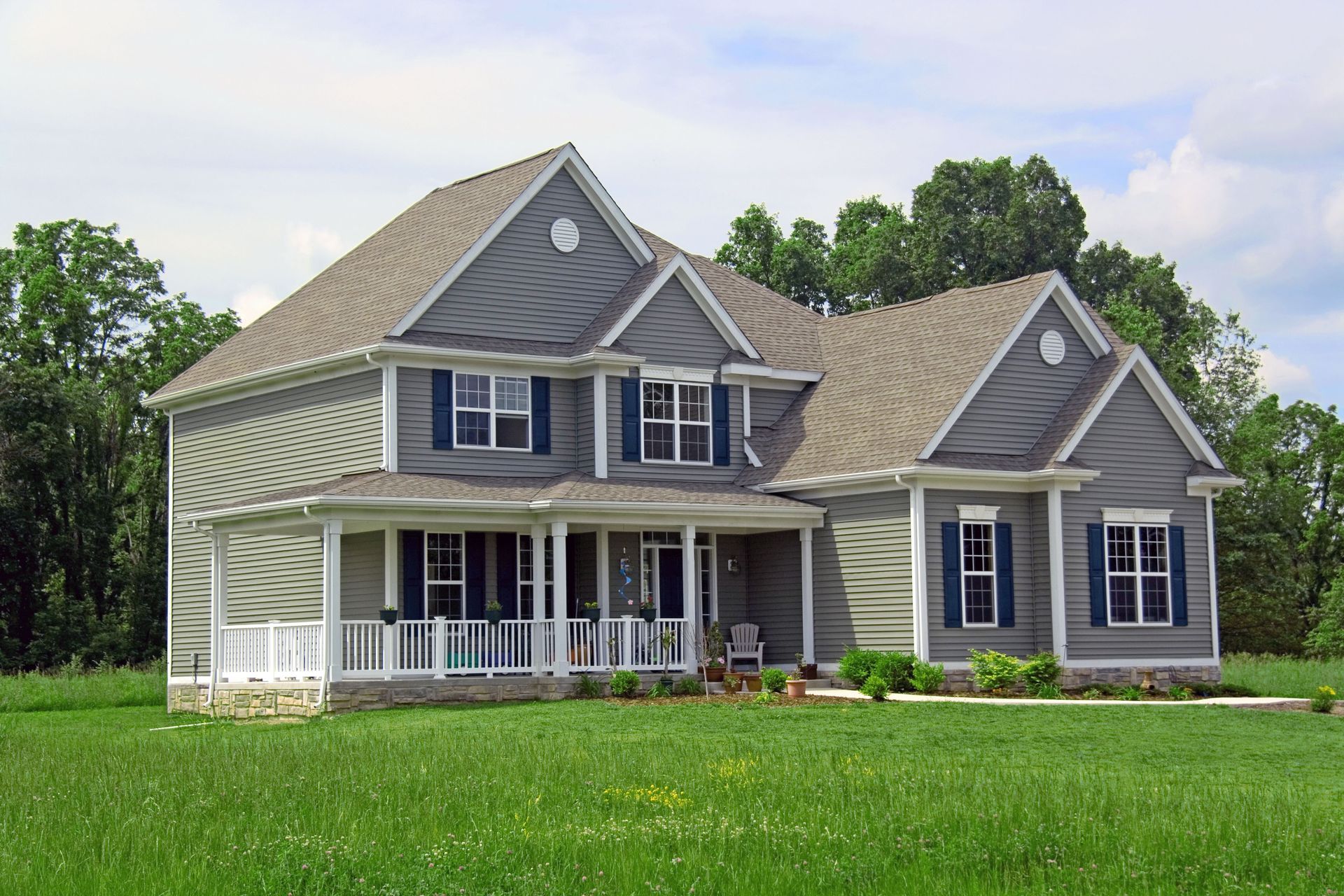Gray two-story house with blue shutters and a wraparound porch, set in a grassy yard under a blue sky.