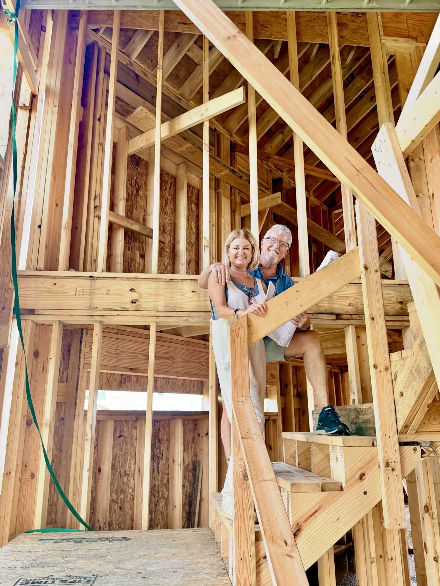 Two people standing inside a wooden house frame under construction, beside unfinished stairs