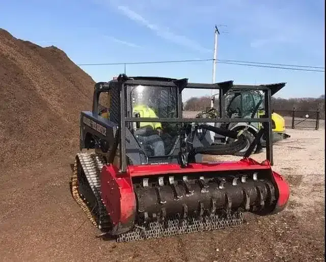 A compact track loader with a red mulcher attachment sits in a dirt lot next to a large pile of mulch under a clear sky.