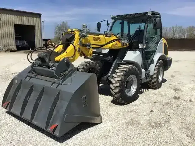 Yellow Wacker Neuson wheel loader with a large grey bucket parked on a gravel lot next to a building.