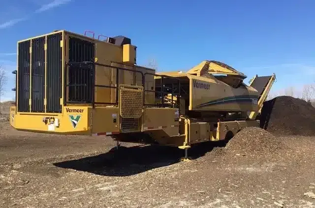A yellow Vermeer horizontal grinder sits on a dirt lot, processing organic material into a pile of mulch.