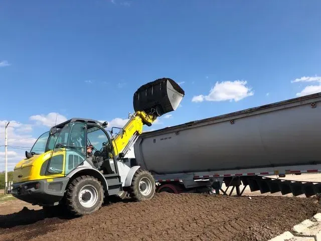 A yellow and gray front-end loader lifts a bucket of soil into the back of a large semi-trailer outdoors.