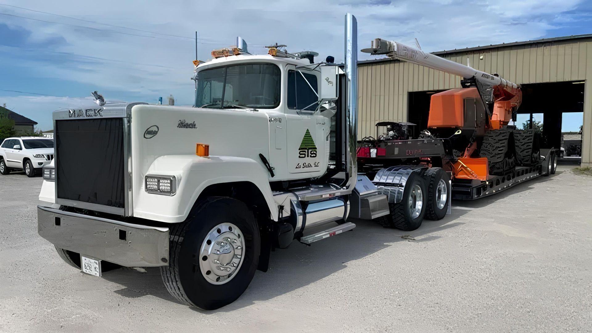 A white Mack semi-truck hauling heavy construction machinery on a flatbed trailer in a gravel lot.
