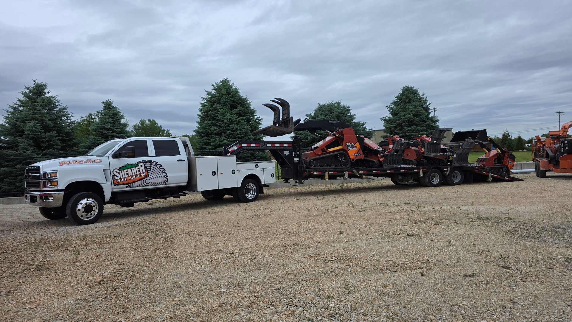 White utility truck pulling orange farm equipment across a gravel lot under cloudy skies