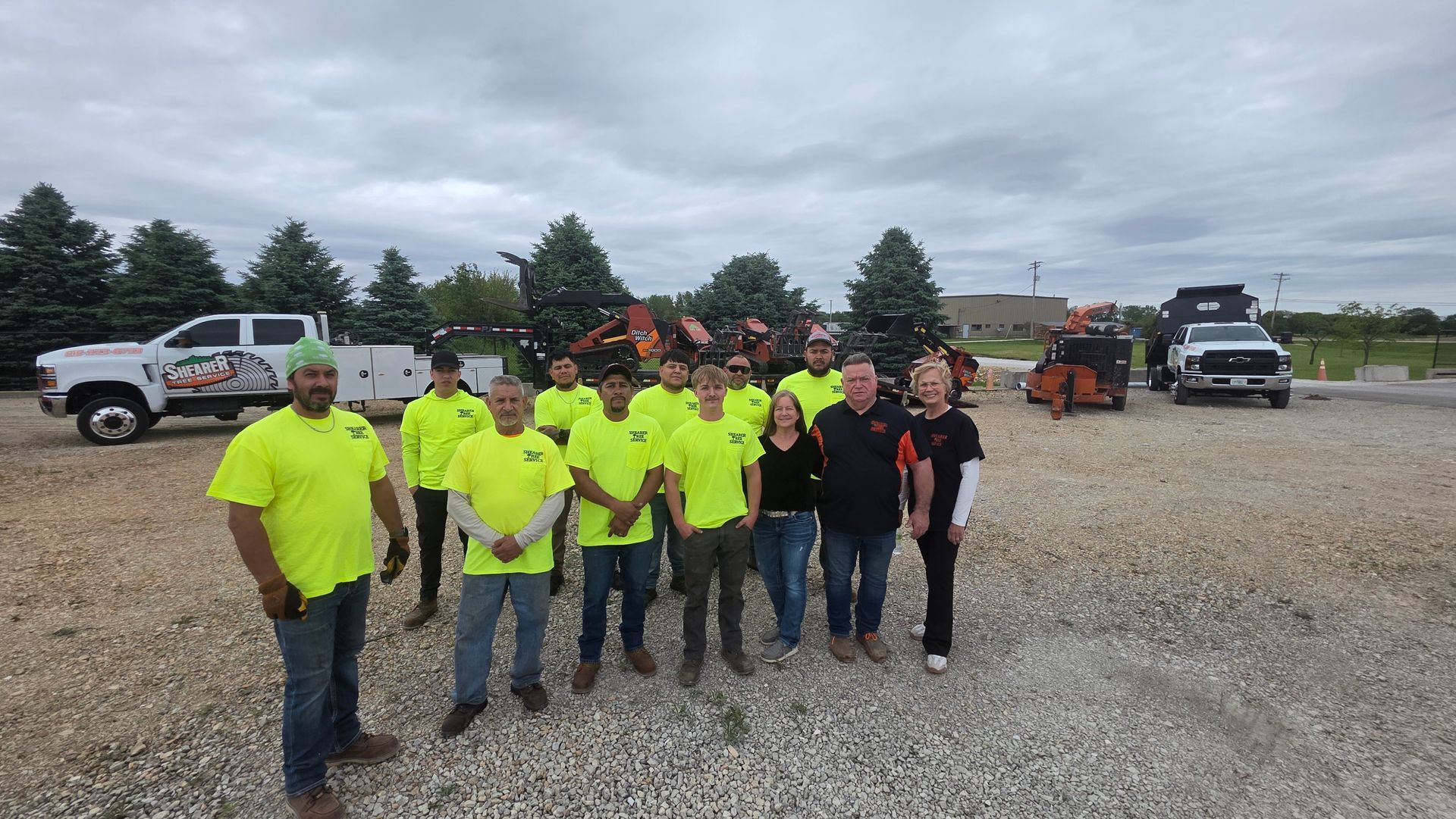 Group of workers in neon shirts and jackets standing on a gravel lot with trucks and equipment behind them
