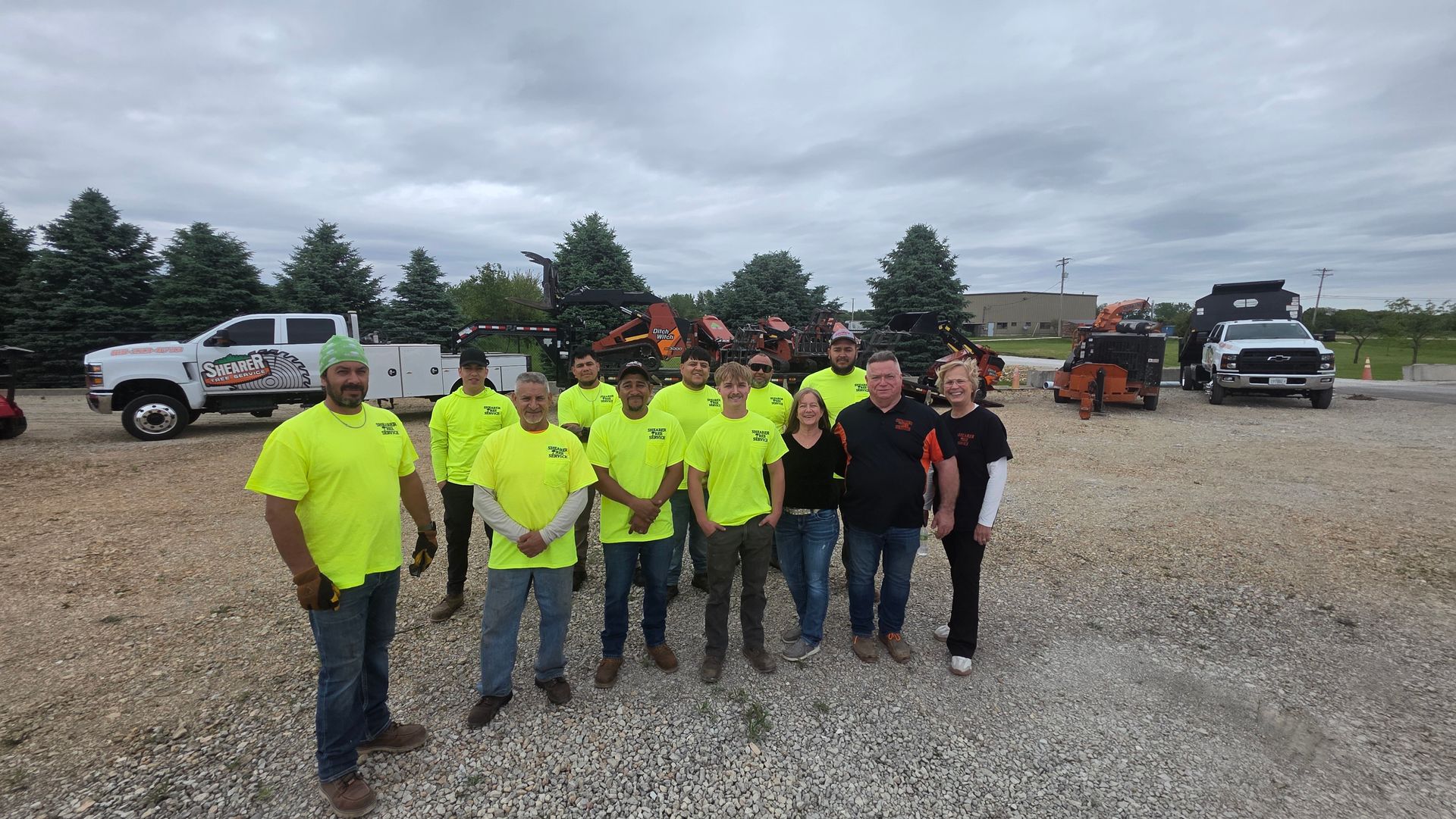 Group of people in neon safety shirts standing on a gravel lot with vehicles and equipment behind them