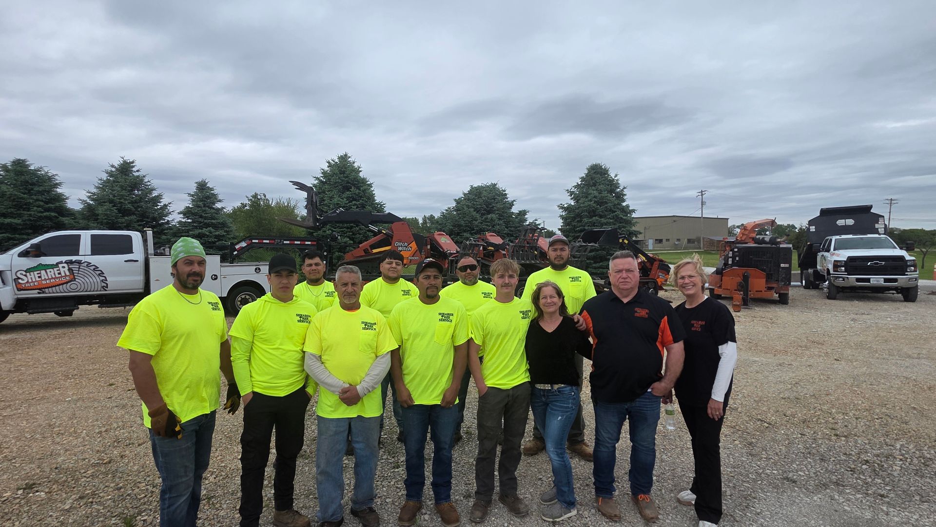 Group of workers in neon shirts and black jackets posing in a gravel lot with trucks and equipment behind them
