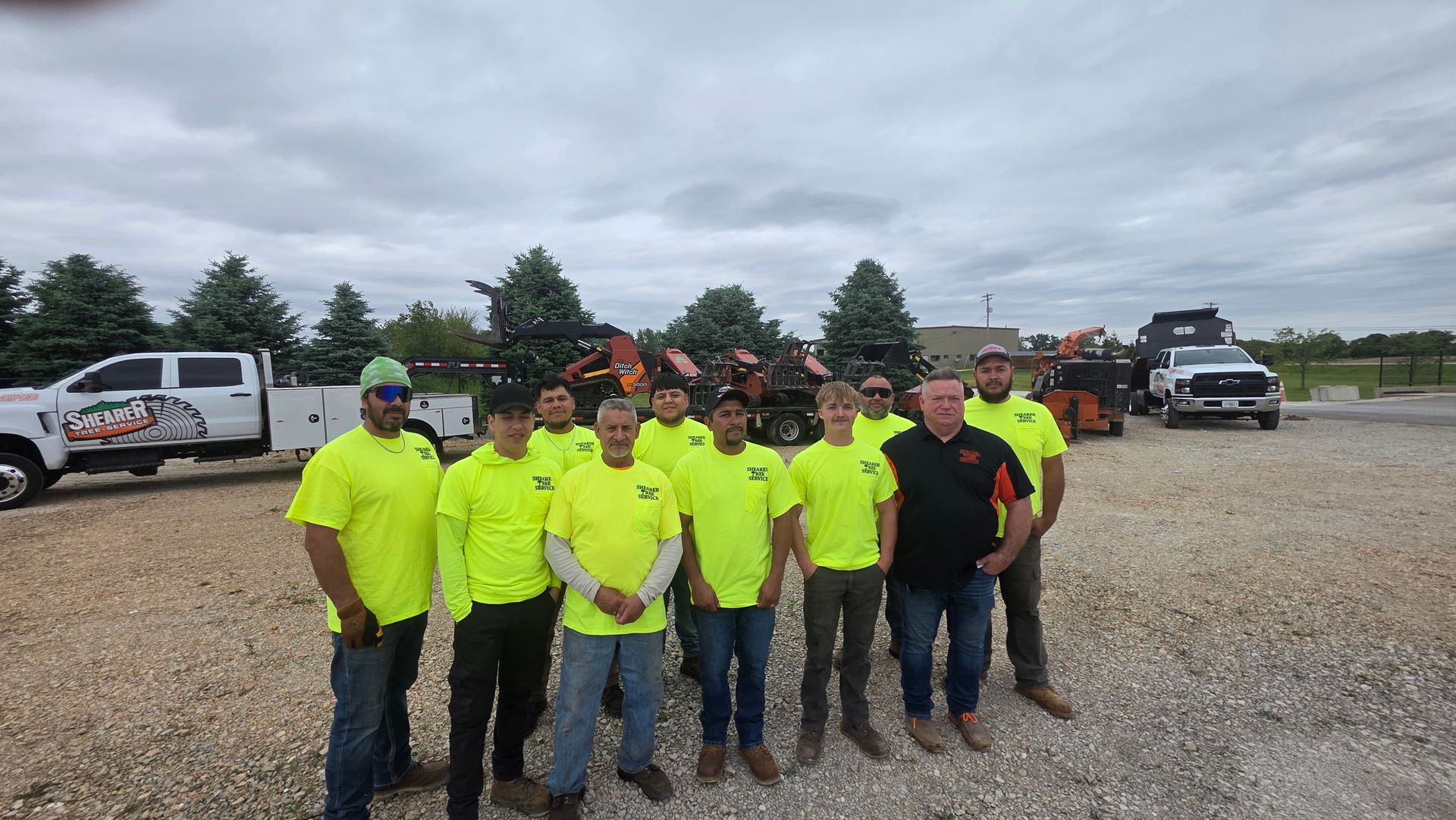 Group of workers in neon safety shirts posing in a gravel lot with trucks and equipment behind them.