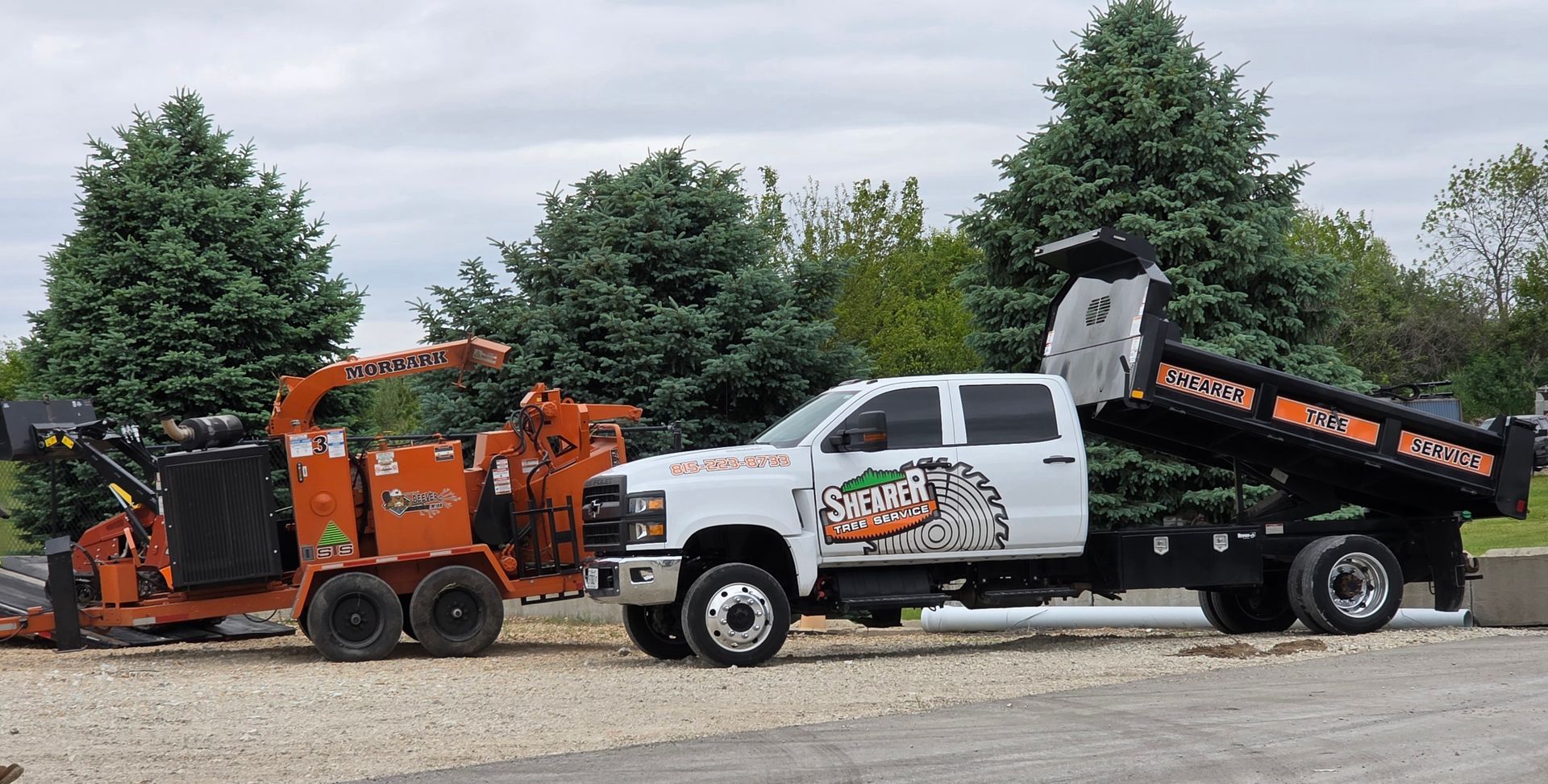 White pickup towing orange wood chipper beside black dump truck in a gravel lot, with trees in back