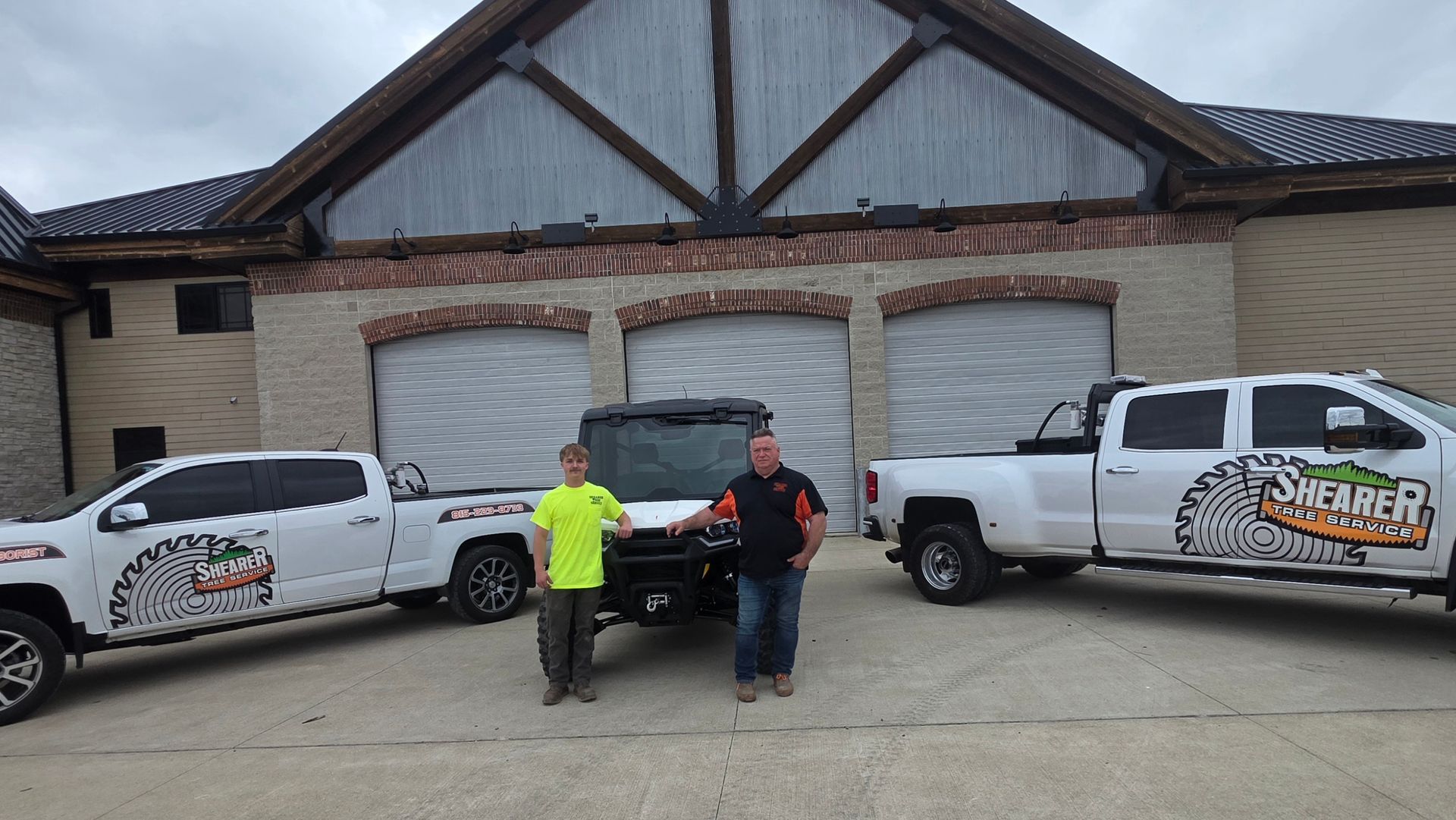 Two men stand between two branded pickup trucks in front of a brick house with a garage.