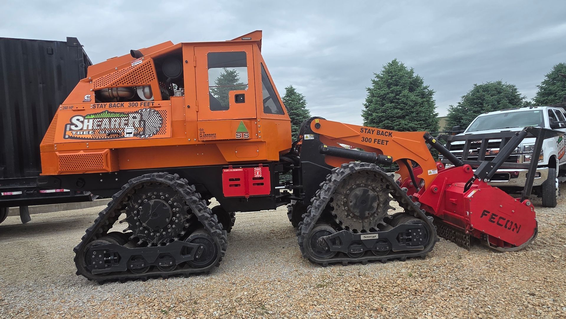 Orange tracked excavator with red grapple attachment parked on gravel beside a black dump trailer