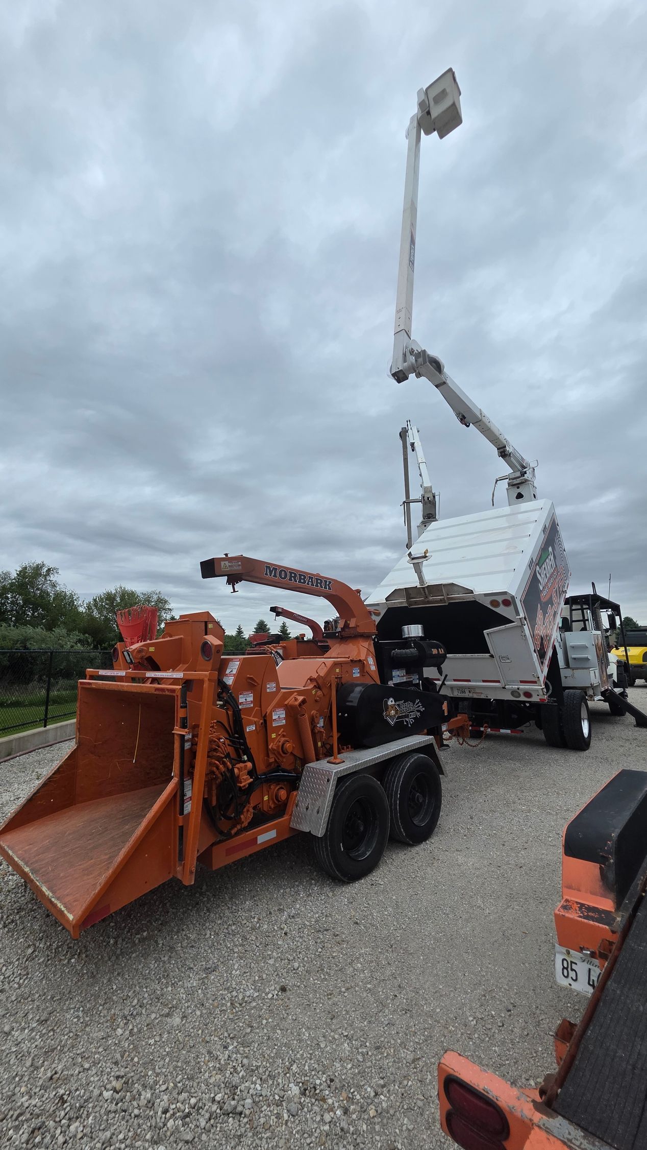 Orange snow blower and a raised utility truck in a gravel lot under cloudy skies