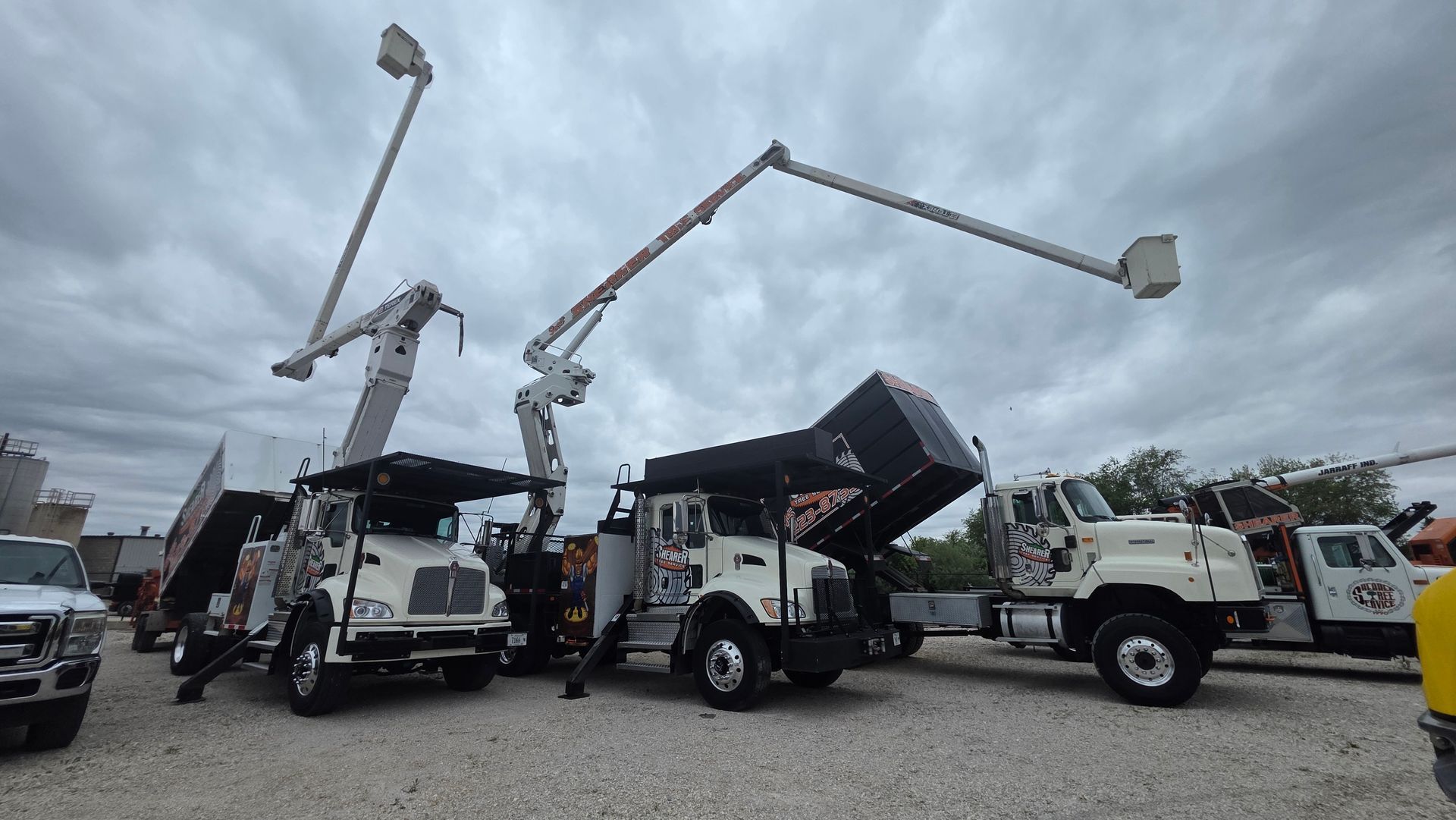 Utility trucks with extended bucket booms lifting a dark dump bed in a gravel lot under cloudy skies