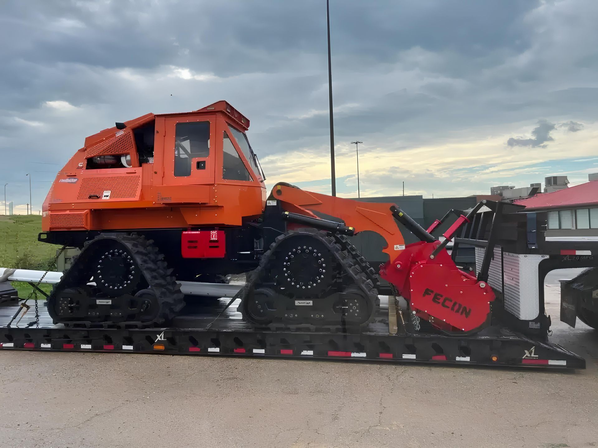 An orange track-loader with a red Fecon mulching head mounted on a flatbed trailer against a cloudy sky.