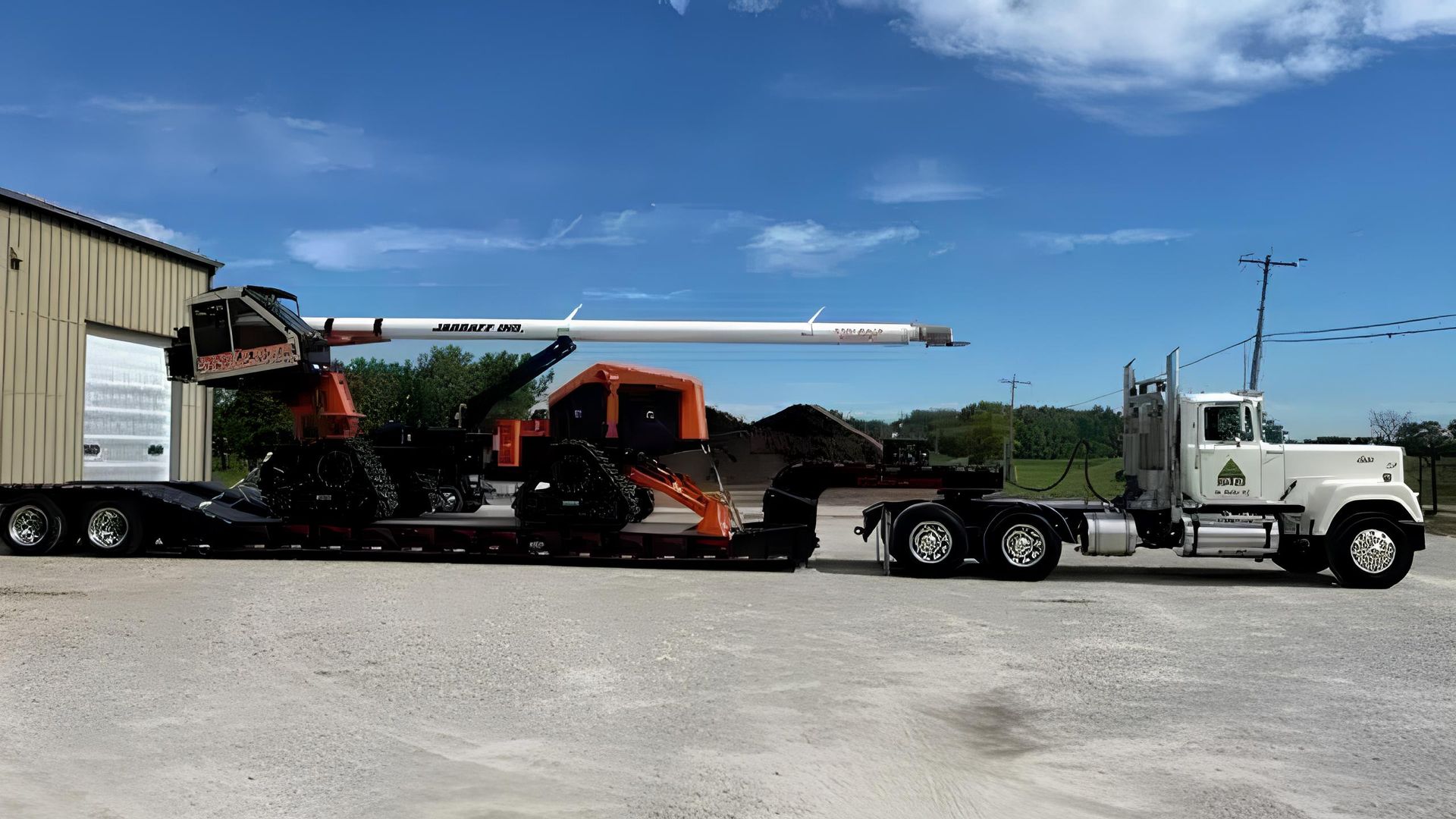 A white semi-truck hauling a flatbed trailer loaded with a piece of orange and black construction equipment.
