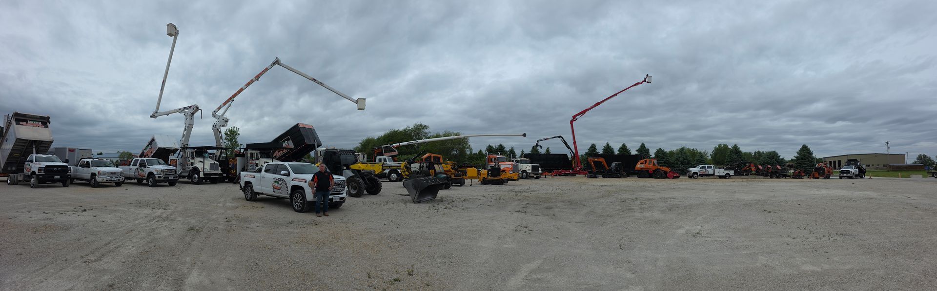 Wide gravel lot with white utility trucks, construction equipment, and a red boom lift under a cloudy sky