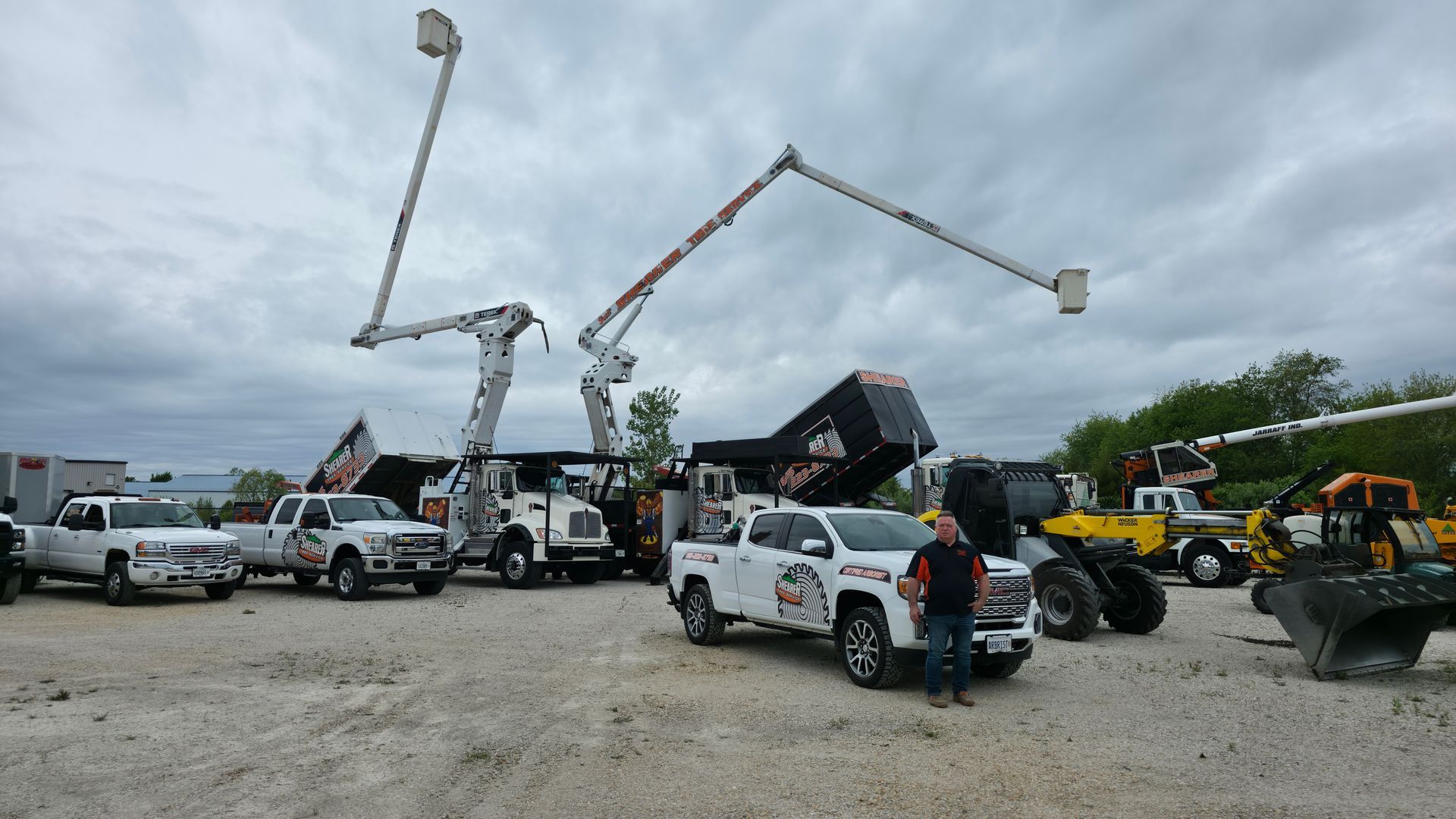 Utility trucks with raised boom lifts and workers in a gravel lot under cloudy skies