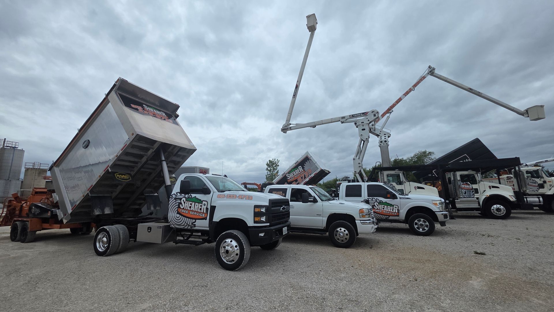 Tow trucks and bucket trucks lined up in a gravel lot under a cloudy sky.