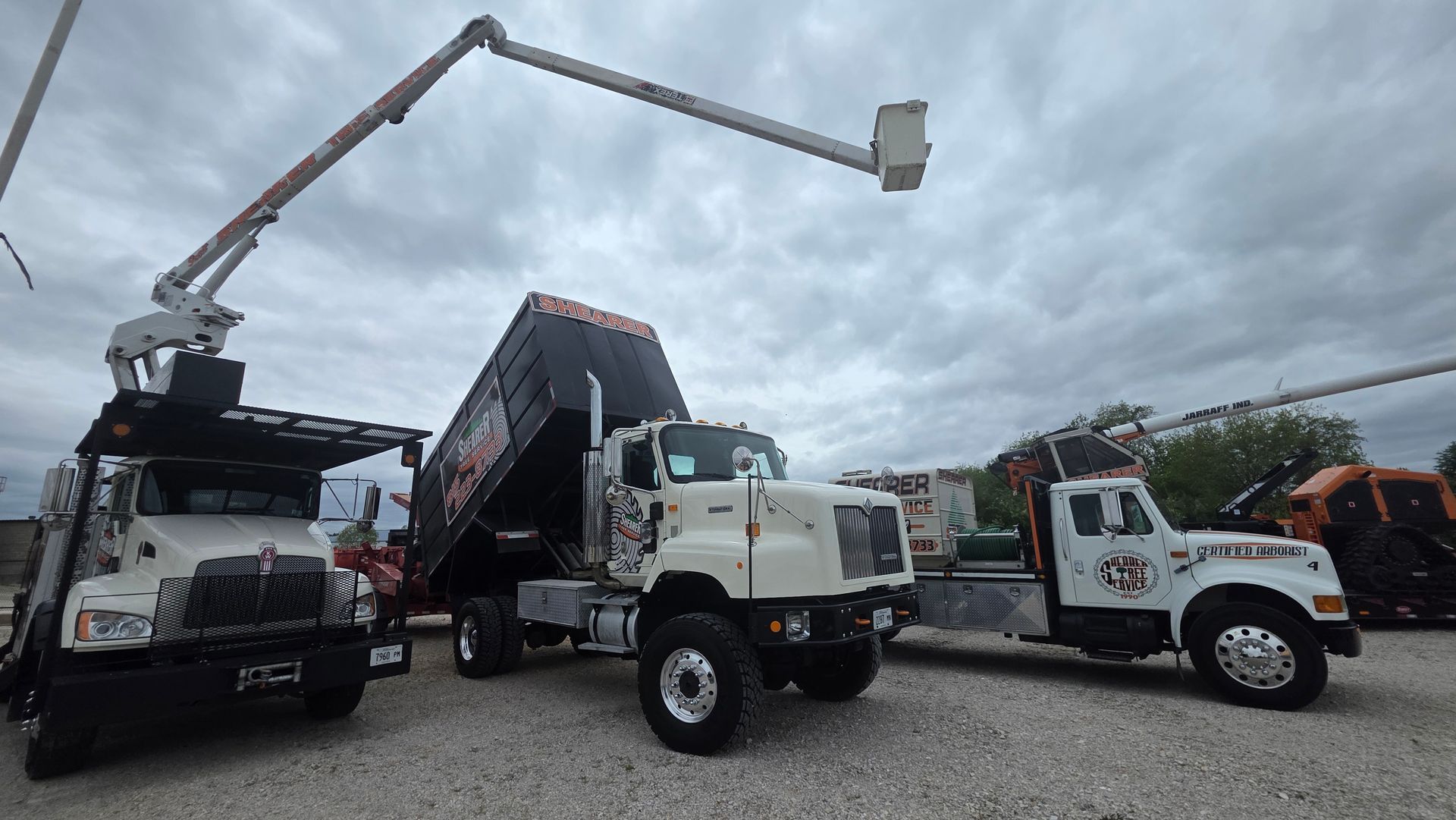 Three utility trucks and an aerial bucket lift parked on gravel under a cloudy sky.