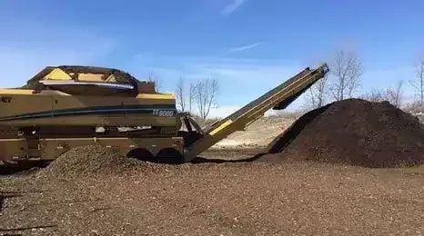 A yellow industrial grinder processing mulch, with a conveyor belt piling material onto a large mound under a blue sky.